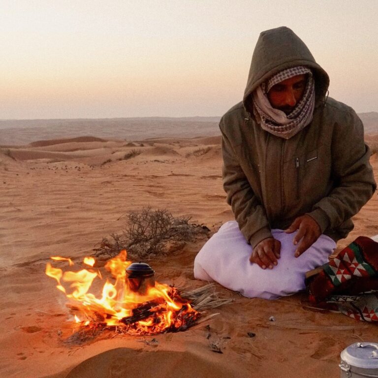 Omani man sitting by a small fire in the Wahiba Sands desert at dawn